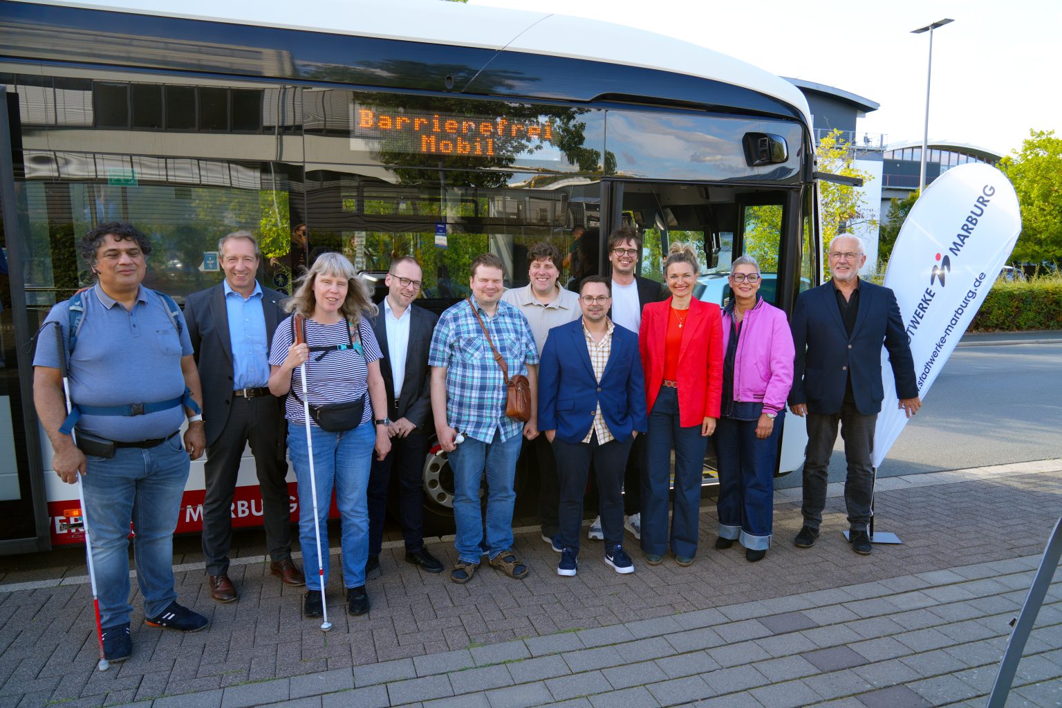 Bürgermeisterin und Stadtwerke-Aufsichtsratsvorsitzende Nadine Bernshausen steht bei der Pressekonferenz zur Vorstellung neuer barrierefreier Fahrpläne gemeinsam mit Vertreterinnen und Vertretern der Stadtwerke Marburg, der blista und OVA Media vor einem Bus. Auf dessen elektronischer Anzeige steht „Barrierefrei Mobil“.